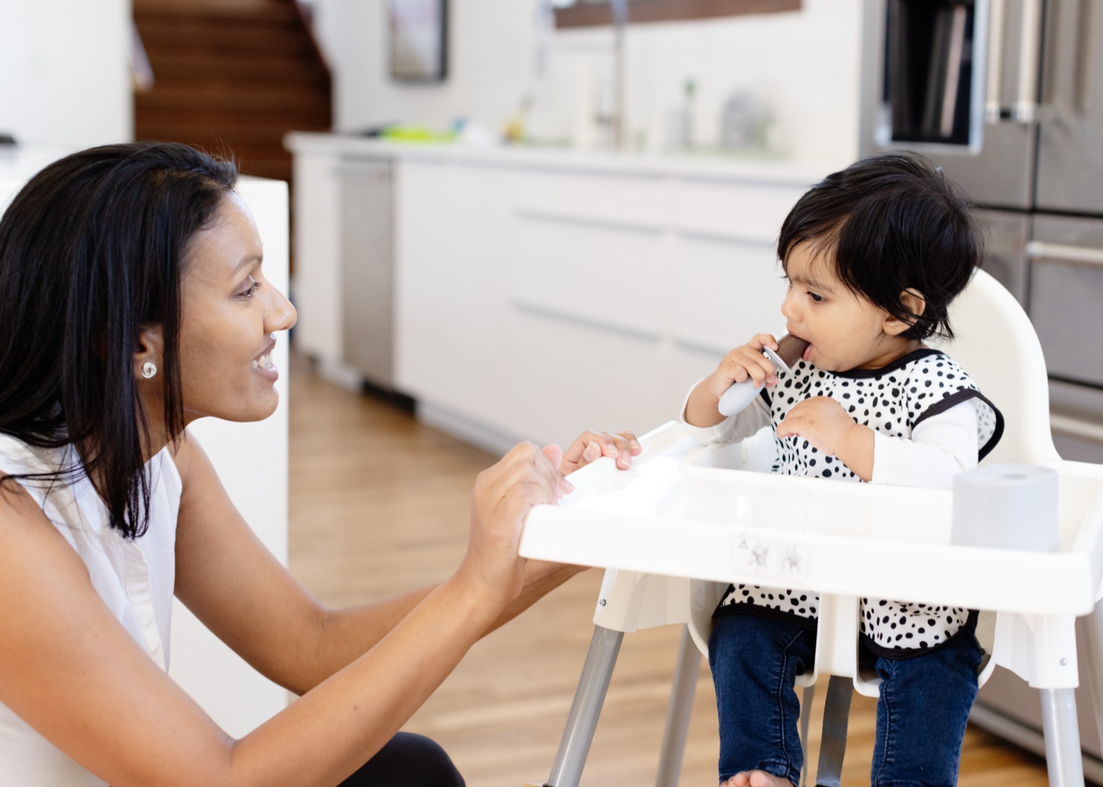 A parent leaning in and talking with her toddler at a table, who points back while eating from a divided plate.